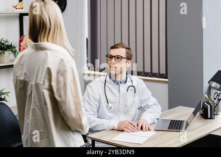 Ein attraktiver junger Arzt hat eine Sprechstunde in seiner Praxis. Der Arzt gibt seinem Patienten Empfehlungen, während er am Arbeitsplatz sitzt. Stockfoto