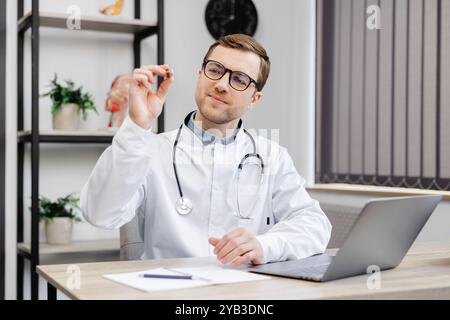 Junger attraktiver Arzt im Hals-Nasen-Ohren-Bereich, der an seinem Arbeitsplatz in der Praxis sitzt und einen Ohrtrichter in den Händen hält. Stockfoto