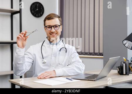 Junger attraktiver Hals-Nasen-Ohren-Arzt, der an seinem Arbeitsplatz in der Praxis sitzt und einen Nasenspiegel in der Hand hält. Stockfoto