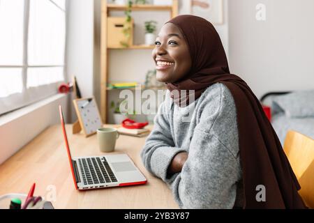 Lächelnde junge afrikanische muslimin mit Laptop, die am Schreibtisch in ihrem Zimmer sitzt Stockfoto