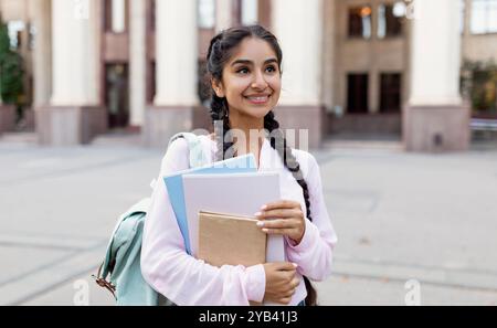 Porträt einer fröhlichen indischen Studentin im Freien mit Rucksack und Arbeitsbüchern, die in der Nähe des College-Gebäudes stehen Stockfoto