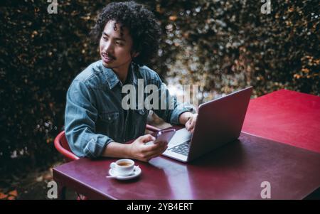 Ein junger asiatischer Mann mit lockigen Haaren sitzt draußen an einem roten Tisch mit einem Laptop und hält ein Smartphone in der Hand. Er ist konzentriert und genießt Kaffee in einem kleinen Stockfoto