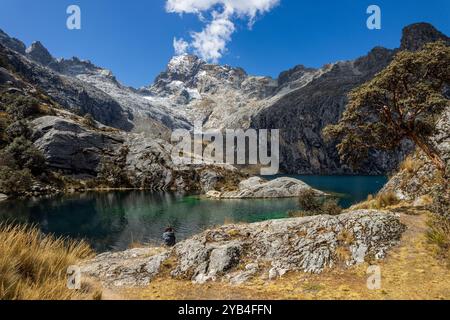 Frau sitzt an der Churup Lagune mit türkisfarbenem Wasser unter dem schneebedeckten Gipfel der Cordillera Blanca im Huascaran Nationalpark in Ancash, per Stockfoto