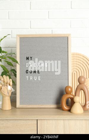 Brieftafel mit dem Satz My Dreams Come True, Pflanzen- und Dekorationselemente auf einem hölzernen Beistelltisch in der Nähe der weißen Ziegelwand Stockfoto