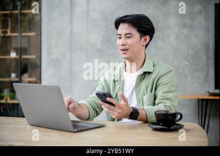 Ein gutaussehender, positiver asiatischer Mann arbeitet aus der Ferne von einem Café aus, hält sein Smartphone in der Hand, während er über seinen Laptop an einem Online-Meeting teilnimmt. Stockfoto