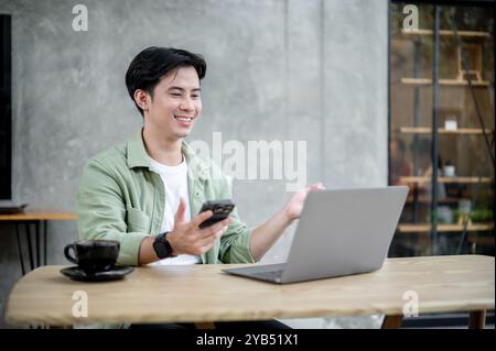 Ein gutaussehender, positiver asiatischer Mann arbeitet aus der Ferne von einem Café aus, hält sein Smartphone in der Hand, während er über seinen Laptop an einem Online-Meeting teilnimmt. Stockfoto