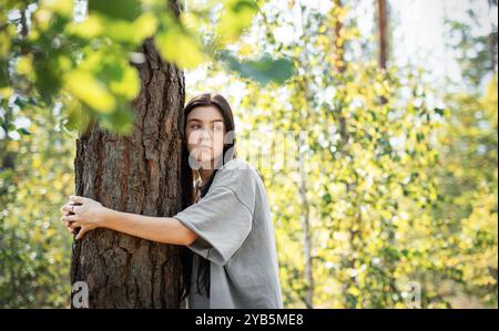 Eine Frau lehnt sich an einen Baum, blickt mit einem ruhigen Ausdruck nach oben, umgeben von leuchtend grünen Blättern in einem sonnendurchfluteten Wald. Stockfoto