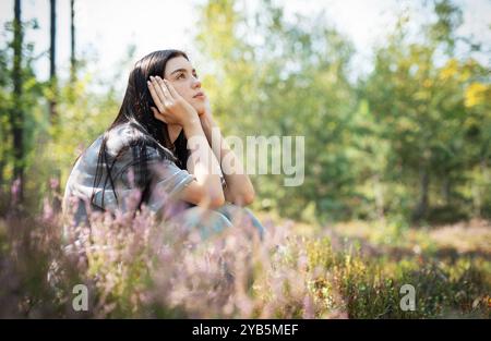 Die junge Frau ist umgeben von blühenden Wildblumen und grünen Bäumen, die in Gedanken verloren sind, während sie sich an einem ruhigen Nachmittag im warmen Sonnenlicht sonnt. Stockfoto