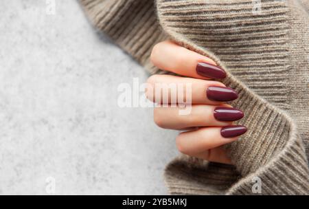 Eine gut gepflegte Hand zeigt dunkle, burgunderrote Nägel und hält einen gemütlichen Strickpullover. Die warmen Töne sorgen für ein einladendes, modisches Ambiente Stockfoto