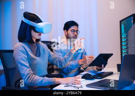 Asiatische Softwareentwickler, die ein Virtual-Reality-Headset tragen, arbeiten an einem VR-Projekt, wobei sich eine Kollegin auf ihren Computer konzentriert Stockfoto