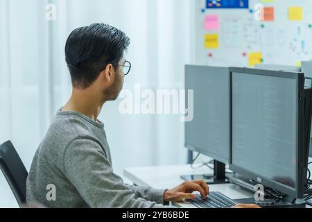 Der Prompt-Ingenieur des asiatischen Mannes entwickelt eine Codierungs-App mit Softwaredaten, die vor dem Computermonitor im Büro sitzen Stockfoto