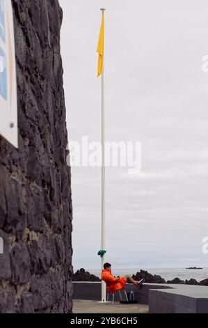 Der Rettungsschwimmer in leuchtendem Orange sitzt unter einer gelben Flagge an den natürlichen Pools von Porto Moniz, neben vulkanischen Steinmauern Stockfoto