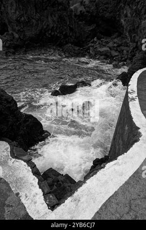 Die Wellen brechen an den natürlichen Pools von Porto Moniz in Schwarz-weiß gegen vulkanische Felsen unter einem scharfen Betonrand Stockfoto