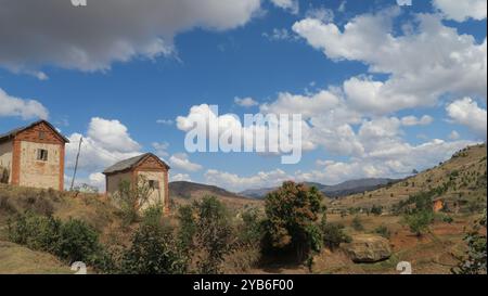 Typische Häuser im Hochland Madagaskars von der Seite mit Wolken und blauem Himmel inmitten hügeliger Felder und Gebirgszug im Hintergrund. Stockfoto