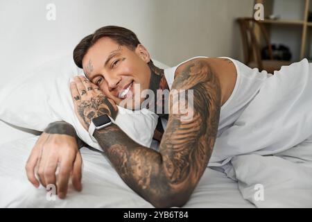 Ein charmanter junger Mann mit Tattoos lächelt, während er zu Hause bequem auf seinem Bett liegt. Stockfoto