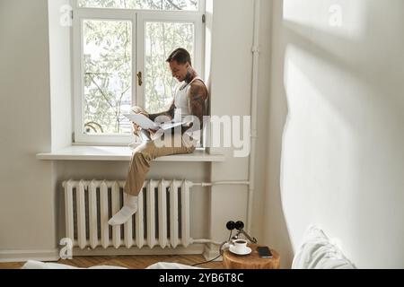 Der junge Mann sitzt auf einer Fensterbank und konzentriert sich auf seinen Laptop, während das Sonnenlicht hereinströmt. Stockfoto