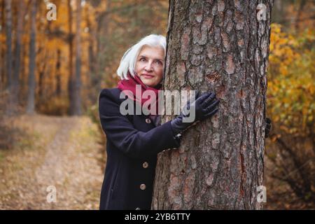 Schöne silberhaarige reife Frau umarmt den Baum im Herbstwald, genießt goldene Blätter fallen, entspannt sich und verbindet sich wieder mit der Natur, friedlich und Stockfoto