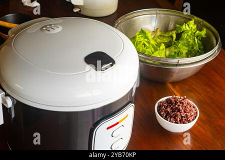 Hochwinkelansicht des elektrischen Reiskochers, gedämpfte braune Reisbeere in weißer Keramikschale und frisches grünes Gemüse im Metallbehälter auf Holztisch. Stockfoto