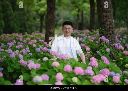 Hübscher asiatischer junger Mann in Brille an bunten Hortensie Makrophylla Blumen, Vorderporträt Stockfoto