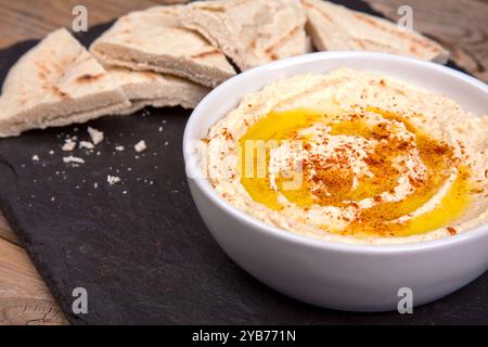Eine Schüssel mit cremigem Hummus, mit Olivenöl bespritzt und mit Paprika bestreut, begleitet von warmen Fladenbrotscheiben, die auf einer dunklen Schieferoberfläche angeordnet sind. Stockfoto