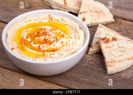 Eine Schüssel mit cremigem Hummus, mit Olivenöl bespritzt und mit Paprika bestreut, begleitet von warmen Fladenbrotscheiben, die auf einer rustikalen Holzoberfläche angeordnet sind Stockfoto
