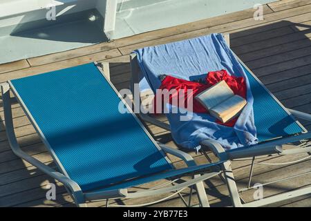 Zwei leere Sonnenliegen auf einer Holzterrasse mit einem Handtuch und einem offenen Buch auf einer. Perfektes Image für Entspannung, Freizeit und Urlaub in Stockfoto