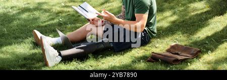 Ein junger Mann mit einer Beinprothese sitzt auf Gras, vertieft in das Lesen eines Buches, umhüllt Natur und Gelassenheit. Stockfoto