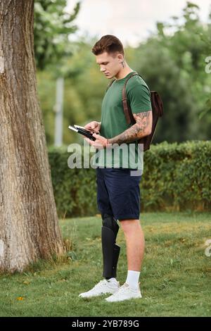 Ein junger Mann mit einer Beinprothese steht in einem üppigen Park und liest sein Buch unter einem Baum. Stockfoto