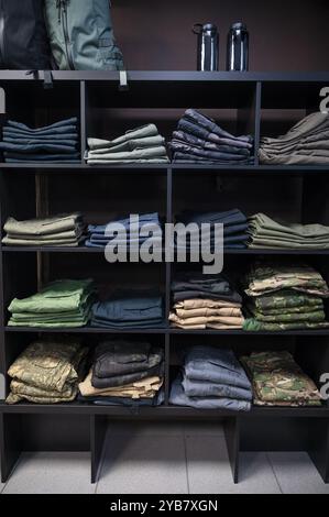 Gestapelte taktische Hosen und Militäruniformen, einschließlich Tarnkleidung und Outdoor-Ausrüstung, ordentlich auf dunklen Holzregalen in einem Geschäft angeordnet. Stockfoto