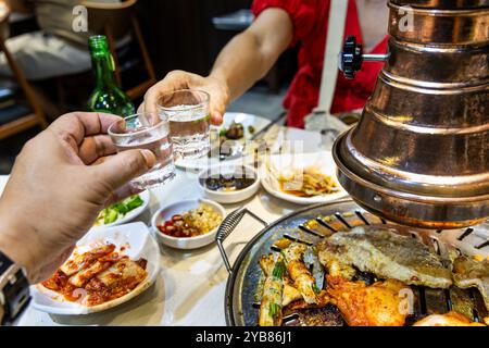 Zwei Personen, die während des Abendessens im koreanischen Grillrestaurant Soju-Getränke anstoßen und dabei Grillfleisch genießen. Stockfoto