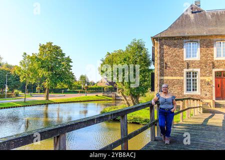 Reife Frau, die auf einer hölzernen Brücke zum Schloss Wijnandsrade steht, lässige Kleidung, Jeans und fröhlicher Ausdruck, Stadtbild im Hintergrund, sonniger Autom Stockfoto