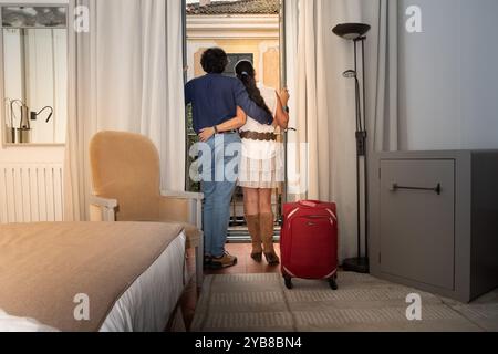 Ein Paar, das mit einem roten Koffer in einem Hotelzimmer mit offenem Fenster abblickte. Von außen sieht man das Gebäude gegenüber, das klassisch EU ist Stockfoto