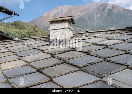 Detail des Schieferdachs mit Schornstein im historischen Bergdorf Valchisone Valley, im hellen Herbstlicht in Usseaux mit Pelvo Peak im Hintergrund Stockfoto