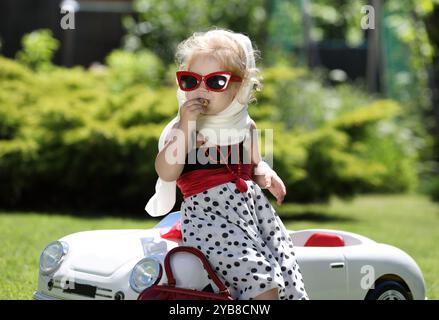 Porträt eines hübschen kleinen blonden Mädchens mit einem weißen Taschentuch, das sich an einem weißen Spielzeugauto im Garten lehnt Stockfoto