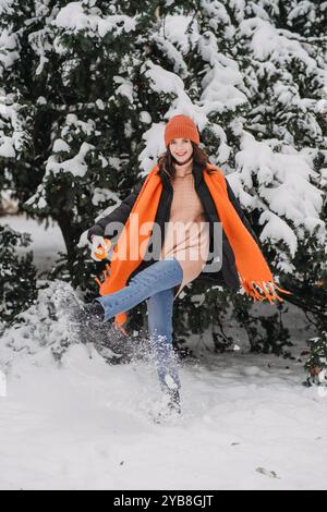 Frau in orangefarbenem Schal und Hut, die im Schnee spaziert, lächelt und den Wintertag im Freien genießt. Wintermodetrends, Naturerlebnisse, Outdoor-Winter Stockfoto