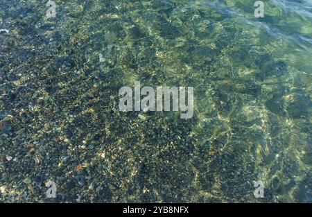 Glitzerndes Sonnenlicht tanzt auf dem Wasser und beleuchtet bunte Kieselsteine, während sanfte Wellen faszinierende Muster an der friedlichen Küste erzeugen. Stockfoto