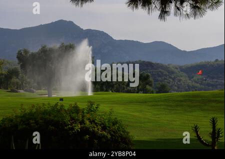 Üppig grüne Fairways erstrecken sich über den Golfplatz, wo ein sanfter Wasserbrunnen im Sonnenlicht tanzt, eingerahmt von majestätischen Bergen und einem Hauch von V Stockfoto