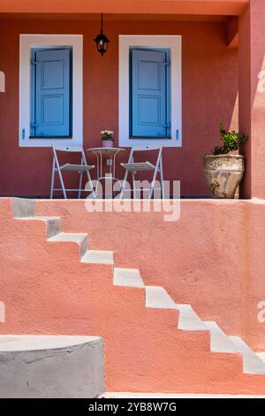 Ein bezauberndes Haus mit rosafarbenen Wänden in Assos, Kefalonia. Die blauen Fensterläden bilden einen markanten Kontrast zur lebendigen Fassade. Eine Terrasse mit einem Tisch und einer Bar Stockfoto