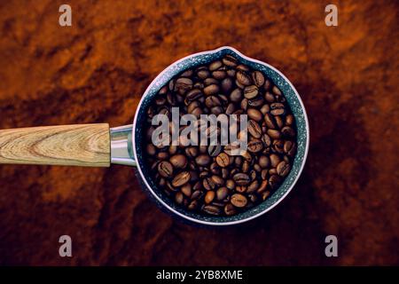 Blick von oben auf eine Kaffeekanne mit Holzgriff. Im Inneren werden geröstete Kaffeebohnen platziert. Die Kaffeekanne steht auf gemahlenem Kaffee. Stockfoto