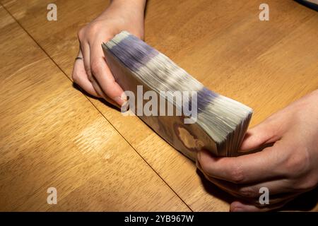 Eine Person, die einen dicken Stapel Bargeld auf einem Holztisch hält. Die Hände befinden sich auf beiden Seiten des Geldes und zeigen die Scheine. Die Ba Stockfoto