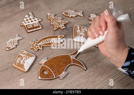 Weibliche Hand, die einen gebackenen Lebkuchenfisch mit süßweißer Zuckerglasur im Gebäckbeutel dekoriert. Nahaufnahme der Zubereitung hausgemachter Weihnachts- und Neujahrskekse. Stockfoto