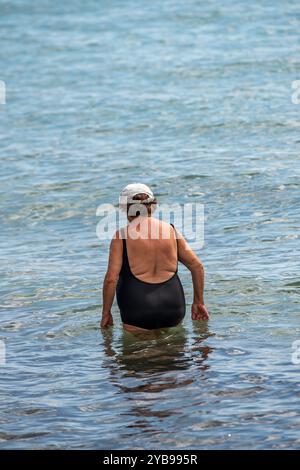 Ältere oder ältere Frau trägt einen schwarzen Badeanzug, der im Meer schwimmt und eine Baseballkappe zum Schutz vor der Sonne trägt. Stockfoto