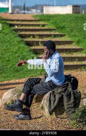 Junger schwarzer Mann, der ein Smartphone benutzt, wird animiert, indem er sich mit Händen während eines Telefongesprächs auf einer Bank ausdrückt. Stockfoto