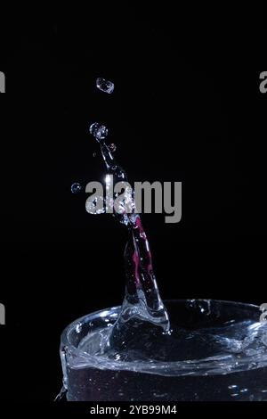 Während des Fluges werden mehrere Wassertropfen erfasst, die aus einem Glasbehälter nach oben spritzen und einen dynamischen Effekt vor einem schwarzen Hintergrund erzeugen. Stockfoto