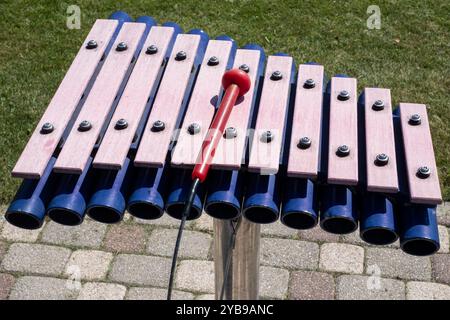 Ein lebhaftes Xylophon steht in einem grasbewachsenen Park mit Holzstangen und farbenfrohen Röhren, die im Sonnenlicht leuchten. In der Nähe erwartet Sie ein markanter roter Hammer Stockfoto