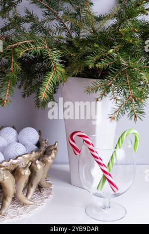 Die saisonale Einrichtung hebt rote und grüne Zuckerstangen hervor, die in einem Glasbehälter platziert werden, gepaart mit leuchtenden immergrünen Zweigen in einer weißen Vase. Stockfoto