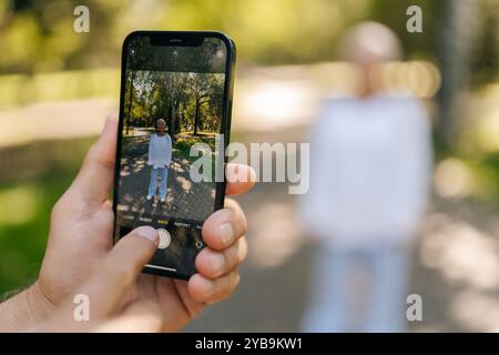 Nahaufnahme einer nicht erkennbaren Seniorin, die sich glücklich im sonnigen Park posiert, während Ehemann Fotograf Momente mit Smartphone festnimmt. Stockfoto