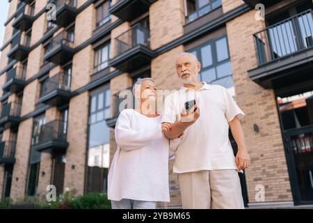 Aufnahmen von unten auf ein fröhliches älteres Paar, das an sonnigem Tag mit einem aussehenden Smartphone vor einem modernen Wohnkomplex aus Ziegelstein steht Stockfoto