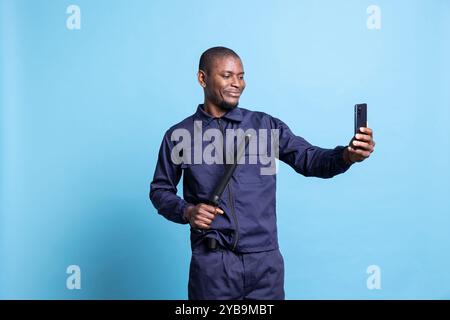 Seriöser Sicherheitsbeamter macht gerne Fotos vor blauem Hintergrund und posiert mit seiner Schlagwaffe auf der Smartphone-Kamera. Selbstbewusster männlicher Wachmann macht Fotos im Studio, Schutzdienst. Stockfoto