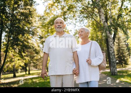 Wunderschönes Seniorenpaar, das einen gemütlichen Spaziergang durch den grünen Park macht, Hände miteinander verflochten hält und an sonnigen Sommertagen einen zärtlichen Moment teilt. Stockfoto